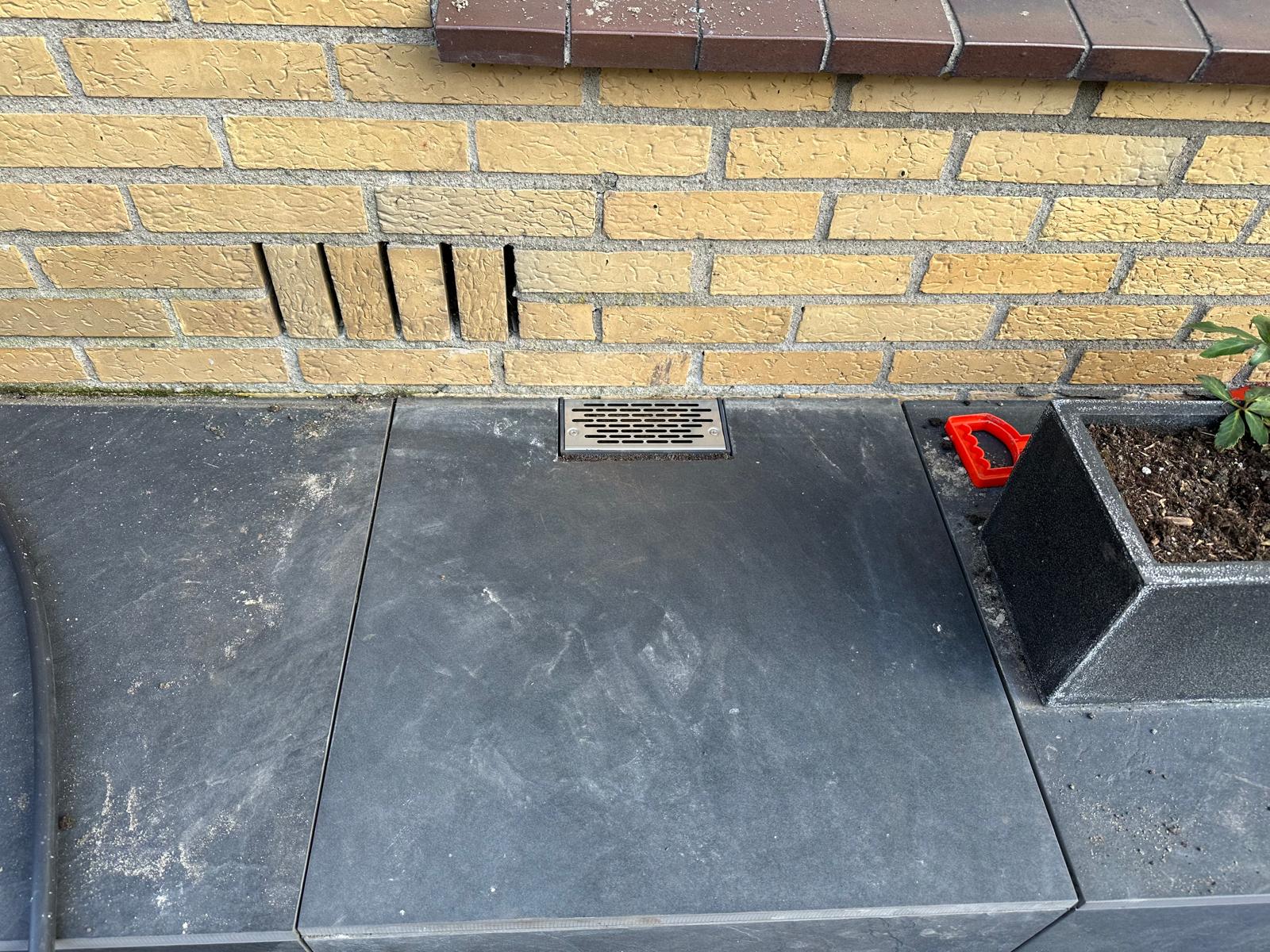 Ventilatierooster in donkere bestrating bij gele baksteenmuur